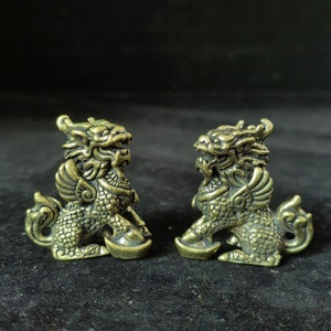 May include: Two antique brass-colored mythical creatures, possibly dragons or lions, are displayed. Each figurine has intricate details, including scales, wings, and a bowl-like object held in its paws. The background is a dark, solid color.