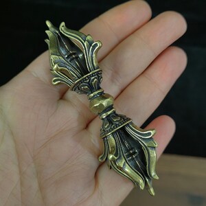 Peut inclure: Un Vajra de couleur bronze détaillé, un objet rituel avec des motifs floraux et géométriques ornés. Les extrémités présentent des pétales stylisés en forme de lotus, et la section centrale a une forme bulbeuse. L'objet est tenu dans une main, mettant en valeur son savoir-faire.