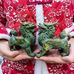 May include: Two bronze-colored mythical lion statues with gold accents. The statues have wings and are held in front of a red and white patterned jacket. The statues are detailed with intricate carvings.