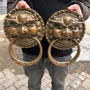 Könnte beinhalten: Zwei antike Türklopfer aus Bronze. Jeder Klopfer zeigt einen detaillierten Löwenkopf mit einem großen Ringgriff. Die Bronze hat eine warme, gealterte Patina. Die Klopfer sind rund und weisen aufwendige Details um das Gesicht des Löwen auf.