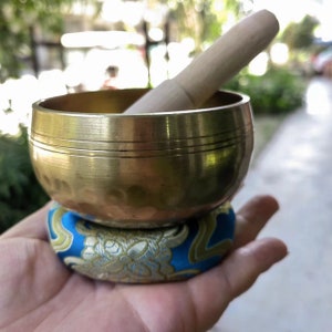 May include: A brass singing bowl with a wooden striker, resting on a blue and gold patterned cushion. The bowl is round with etched lines, and the striker is light-colored wood. The cushion has a floral design.