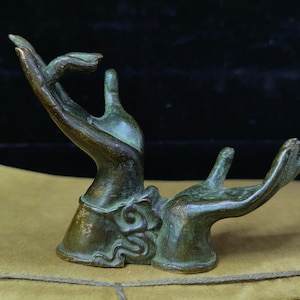 May include: A bronze sculpture of two hands in a gesture of offering or receiving. The hands are detailed with visible knuckles and veins, and have a weathered, aged appearance. The sculpture has a dark green patina, suggesting age and exposure to the elements.