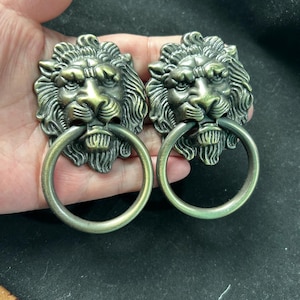 May include: Two antique brass-toned metal lion head door knockers. Each piece features a detailed lion face with a flowing mane and a circular ring held in the lion's mouth. The finish has a darkened, weathered patina with highlights on the raised areas.