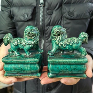 May include: Two matching ceramic Foo Dog statues in a vibrant teal glaze. Each dog is perched atop a square base, with detailed features and a crackle finish. The statues are held in a person's hands, showcasing their size and craftsmanship.