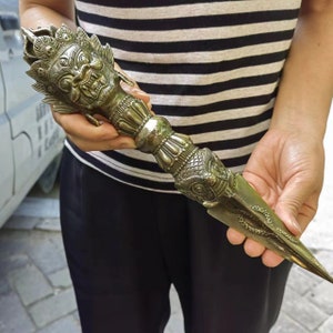 May include: A metallic phurba ritual dagger featuring an intricate three-faced deity head at the pommel, a central lotus-shaped grip, and a pointed blade adorned with a coiled snake relief. The object has a weathered bronze finish with detailed carvings.