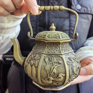 May include: A brass-coloured metal teapot with a hexagonal body and a swing handle. The surface features intricate embossed relief carvings of aquatic plants, fish, and birds. It includes a matching lid with a small knob and a curved spout.