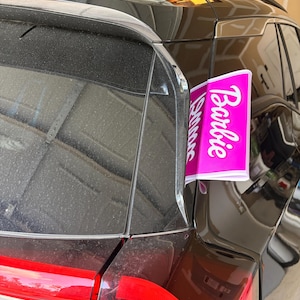 May include: A bright pink rectangular car flag featuring the white text Barbie printed vertically. The flag is attached to the rear window frame of a black vehicle, positioned near the red taillight and the black roof rail of the car.