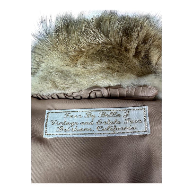 May include: Close-up of a vintage fur coat collar with a light brown fur trim. The coat's lining is a light tan color, and a label reads "Furs By Bella F Vintage and Estate Furs Brisbane, California."