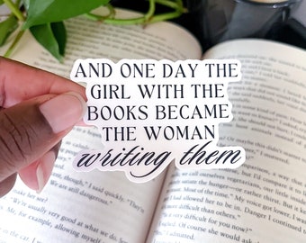 May include: A white sticker with black text that reads "And one day the girl with the books became the woman writing them". The sticker is placed on a book with a white background.