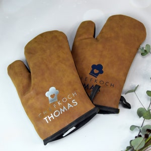 Könnte beinhalten: Zwei braune Ofenhandschuhe mit schwarzem Besatz. Ein Handschuh hat eine silberne Kochmütze mit dem Text "Chef Koch Thomas" und der andere Handschuh hat eine blaue Kochmütze mit dem Text "Chef Koch Max".
