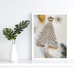 以下が含まれることがあります： 白いマクラメのクリスマスツリーの壁掛けで、木製の星が頂上にあります。ツリーは白い壁に白いフレームで囲まれた状態で吊り下げられています。