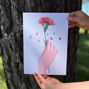 May include: A print featuring a pink carnation with miniature figures on swings suspended from the flower's petals. A stylized pink hand holds the flower's stem. The artwork has a whimsical and surreal quality.