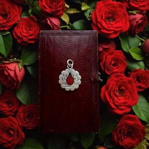 May include: A silver charm featuring a red ladybird design is attached to a dark red, rectangular object. The object is surrounded by vibrant red roses and green leaves, creating a romantic and natural aesthetic.