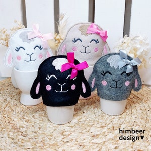 May include: Three felt lamb egg cozies in white, black, and gray. Each lamb has a pink or gray bow and embroidered eyes and a nose. The cozies are sitting in white egg cups.