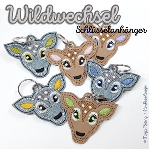 以下が含まれることがあります： 鹿の顔のデザインが施された、5種類の異なる色の革製のキーホルダー。キーホルダーは、茶色、灰色、ベージュで、異なる色のアクセントが付けられています。画像の右上隅には、「Wildwechsel Schlüsselanhänger」というテキストが表示されています。