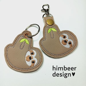 Puede incluir: Dos llaveros de cuero marrón con perezosos bordados colgando de anillos metálicos. Los perezosos están colgando boca abajo con hojas verdes. El texto "himbeer design" está impreso debajo de los llaveros.