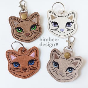 May include: Four cat-shaped keyrings in shades of brown, white, tan, and reddish-brown. Each keyring features a cat face with embroidered details, including eyes, whiskers, and a nose. The keyrings have metal clasps and key rings. The text "himbeer design" is visible.