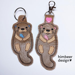 Könnte beinhalten: Zwei braune Otter-Schlüsselanhänger mit gestickten Details. Einer hält einen blauen Fisch, der andere einen rosa Fisch. Einer hat einen silbernen Schlüsselring, der andere einen silbernen Karabiner. Der Text "himbeer design" steht unten rechts.