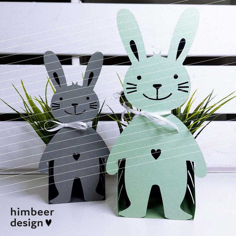 May include: Two papercraft bunny decorations, one gray and one green, with black eyes and a black heart on their chests. The bunnies are standing on a white surface with green grass behind them.