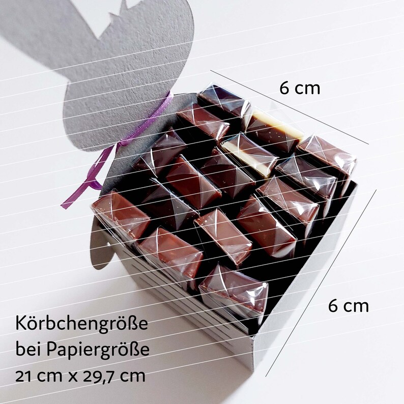 May include: A gray cardboard bunny basket filled with individually wrapped chocolate squares. The basket measures 6 inches by 6 inches and is tied with a purple ribbon. The text on the basket reads "K&ouml;rbchengr&ouml;&szlig;e bei Papiergr&ouml;&szlig;e 21 cm x 29,7 cm".