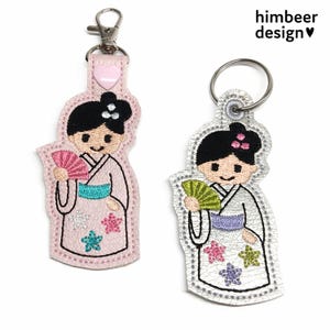 Pode incluir: Dois chaveiros com um design de gueixa japonesa. Um é rosa com um mosquetão, o outro é prateado com um porta-chaves. Ambos apresentam uma gueixa segurando um leque, com detalhes florais. O texto "himbeer design" é visível.