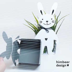 May include: White bunny planter with a black bow and a small black heart on the front. The planter is made of metal and has a cutout design. The bunny is holding a small green plant in its arms. The planter is sitting on a white surface. The text "himbeer design" is printed on the surface below the planter.