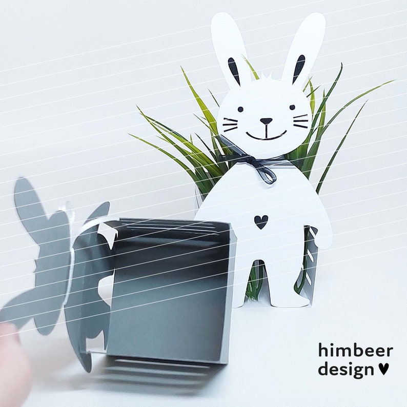 May include: White bunny planter with a black bow and a small black heart on the front. The planter is made of metal and has a cutout design. The bunny is holding a small green plant in its arms. The planter is sitting on a white surface. The text "himbeer design" is printed on the surface below the planter.