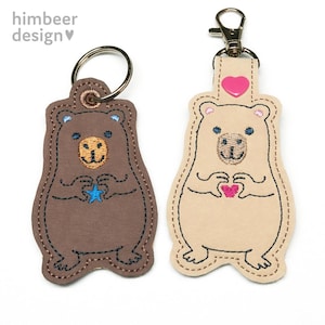 May include: Two bear-shaped keychains. One is brown with a blue star, the other is tan with a pink heart. Both have embroidered details, including eyes, noses, and outlines. The keychains have metal rings and clasps.