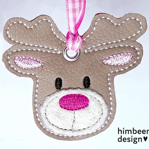 以下が含まれることがあります： 茶色の革のような質感、白いステッチ、ピンクのアクセントが施されたトナカイ型のオーナメント。トナカイは黒い目、ピンクの内側の耳、ピンクの鼻をしています。ピンクと白のストライプのリボンが上部に付いています。「himbeer design」の文字が見えます。