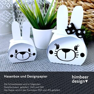 May include: Two white paper bunny boxes with black facial features and upright ears. One box has a black bow with white stars. The boxes are on a gray surface with a small potted plant in the background.