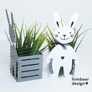 May include: Two white bunny shaped planters with a gray planter in the middle. The planters are filled with green grass. The bunny planters have black eyes, a black nose, and a black mouth. The bunny planters have a black bow tie. The bunny planters have a black heart on their chest. The gray planter has a white heart on its side. The planters are on a white background. The text "himbeer design" is in the bottom right corner of the image.