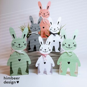May include: Five bunny-shaped paper boxes in different colors, including pink, gray, and green. The boxes are decorated with black eyes and a heart shape. The boxes are arranged in a row on a white surface.