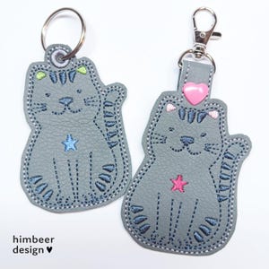 May include: Two gray felt cat keychains with embroidered details. One keychain has a pink heart and a blue star, the other has a green star. Both have a blue embroidered outline.