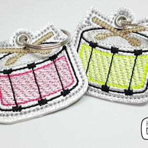 May include: Two embroidered keychains shaped like drums. One drum is pink and the other is green. Both drums have black and white details and gold-colored drumsticks.
