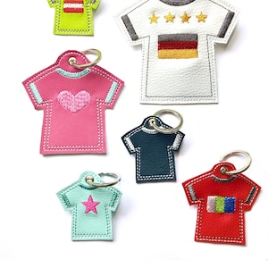 Könnte beinhalten: Sechs bunte Leder-Schlüsselanhänger in T-Shirt-Form mit verschiedenen Stickereien. Die Schlüsselanhänger liegen auf weißem Untergrund. Der Text "himbeer design ♥" befindet sich am unteren Bildrand.