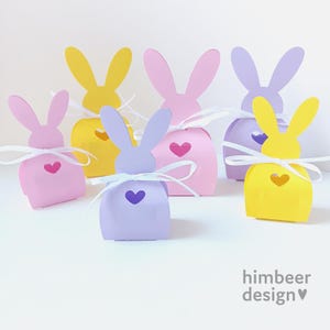 May include: A collection of colorful, bunny-shaped gift boxes in shades of yellow, pink, and purple. Each box features a heart-shaped cutout and is tied with a white ribbon. The boxes are arranged on a white surface, perfect for Easter or spring-themed gifts.