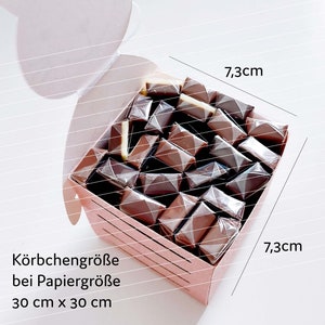 May include: A pink cardboard box filled with individually wrapped chocolate squares. The box measures 7.3 inches by 7.3 inches. The text on the box reads "K&ouml;rbchengr&ouml;&szlig;e bei Papiergr&ouml;&szlig;e 30 cm x 30 cm".