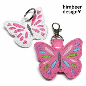 Może przedstawiać: Dwa breloczki do kluczy w kształcie motyla. Jeden jest biały z różowymi akcentami i srebrnym kółkiem na klucze. Drugi jest różowy z niebieskimi, zielonymi i fioletowymi akcentami i srebrnym zapięciem. Widoczny jest tekst "himbeer design".