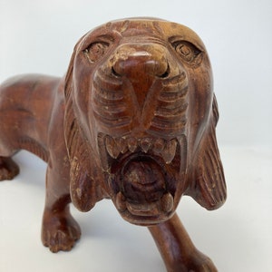 May include: A hand-carved wooden lion sculpture with a snarling expression. The lion is depicted in a walking pose with its head turned to the side. The sculpture is made of dark brown wood and has a rustic finish.
