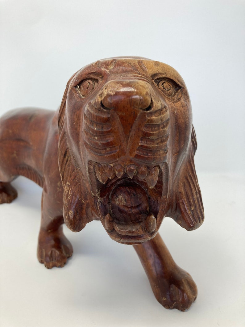 May include: A hand-carved wooden lion sculpture with a snarling expression. The lion is depicted in a walking pose with its head turned to the side. The sculpture is made of dark brown wood and has a rustic finish.