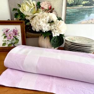 May include: A folded lavender and white striped bed sheet. The sheet is made of a soft fabric and is folded neatly. The background includes a vase of white flowers, a framed floral print, and a stack of plates.
