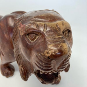 May include: A carved wooden tiger figurine with a detailed face and open mouth. The tiger is brown with a reddish-brown patina.