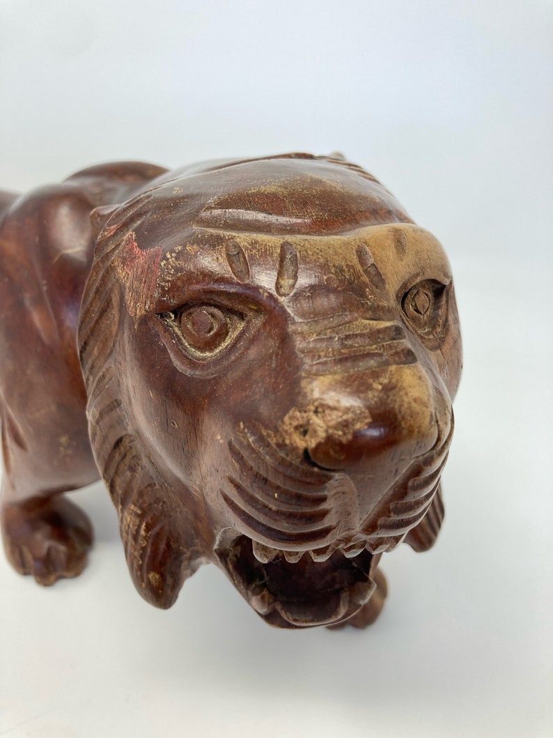 May include: A carved wooden tiger figurine with a detailed face and open mouth. The tiger is brown with a reddish-brown patina.