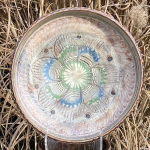 May include: A large, round, white ceramic plate with a decorative design in shades of blue, green, and brown. The design features a central flower with swirling patterns and leaves.