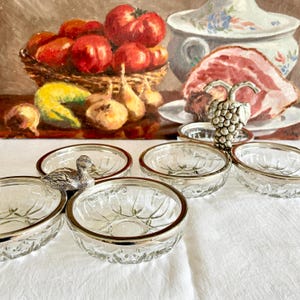 Peut inclure: Ensemble de bols en verre vintage avec des bords argentés. Une figurine décorative de canard argenté et une figurine de grappe de raisin sont également présentes. L'arrière-plan présente une nature morte avec des tomates, des oignons et du jambon.