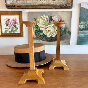 May include: Two wooden candle holders with a light brown finish. Each holder has a round top, a tall, rectangular stem, and a decorative base. A straw hat with a black band sits in the background, along with framed artwork.