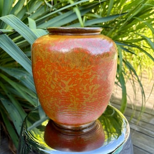 Peut inclure: Un vase en céramique avec un corps large et arrondi et un col étroit. Le vase est principalement orange avec un motif moucheté d'or et de brun. Des rainures horizontales entourent le corps. Le bord et la base sont brun foncé.