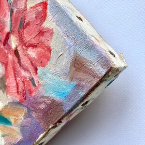May include: Close-up of a colorful abstract painting on a stretched canvas. The painting features a pink flower with blue and white accents. The canvas is secured with staples and has a textured surface.