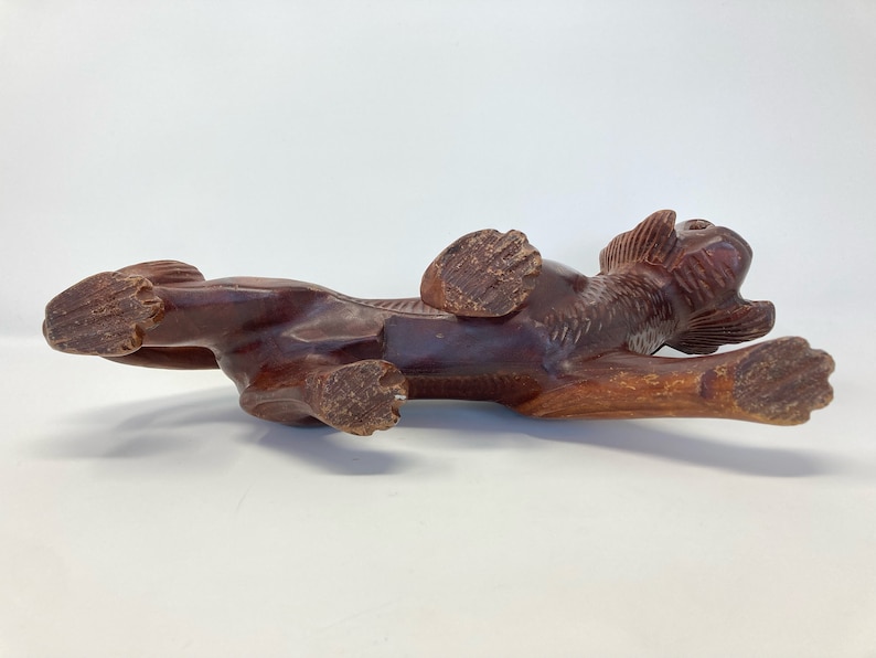 May include: A hand-carved wooden sculpture of a brown lion in a crouching position. The lion's head is turned to the side and its mouth is open. The sculpture is detailed with carved fur and claws.