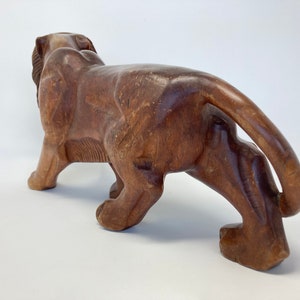 May include: A hand-carved wooden lion figurine with a brown finish. The lion is depicted in a walking pose with its tail curled behind it.